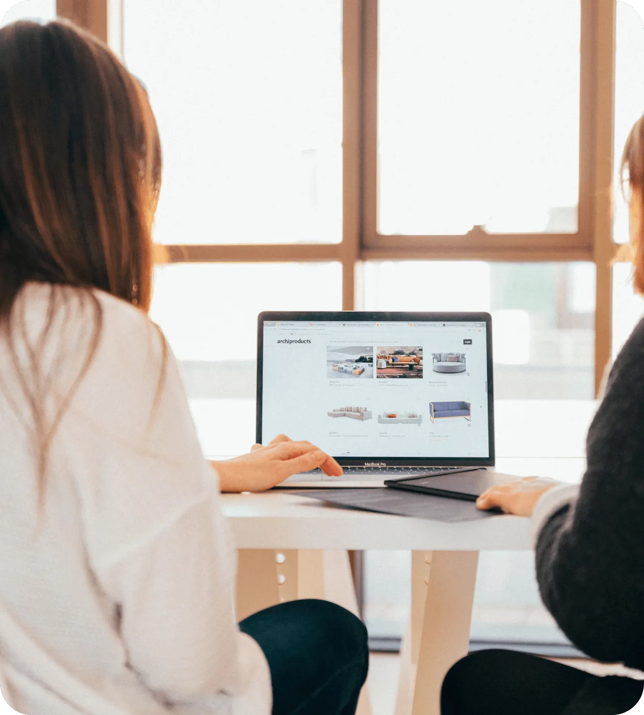 Two people at a table by a bright window looking at a laptop showing a furniture marketplace site with a grid of sofa listings