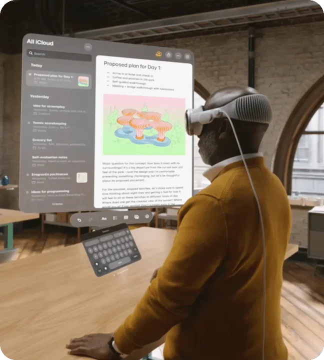 Man in a white spatial-computing headset stands at a table in a brick loft, a large floating notes window and an on-screen keyboard hovering in front of him