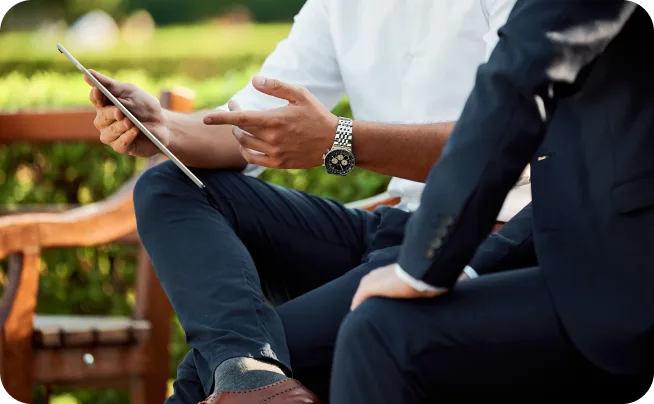 Two men in business attire talking on an outdoor bench, one holding a tablet and stylus