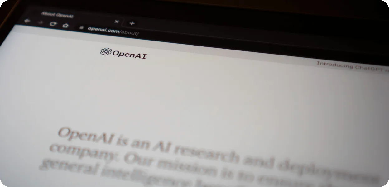 Close-up of a screen showing the About page of AI developer OpenAI, with its knot logo and a headline calling it an AI research and deployment company