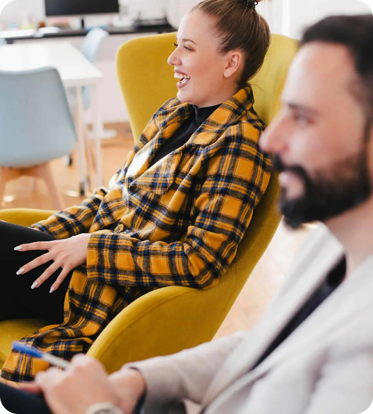 A woman laughing in a yellow armchair during a relaxed conversation, with a colleague in the foreground