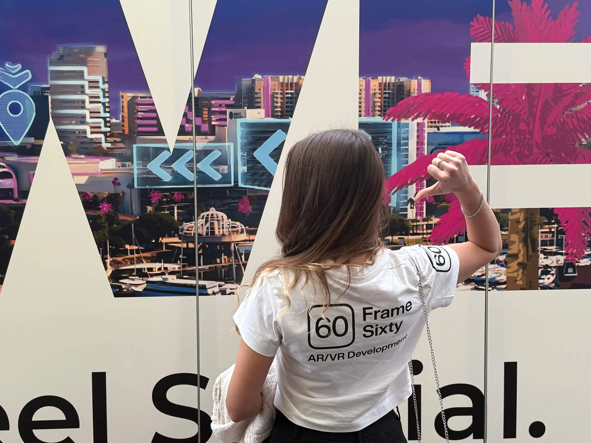 Woman in a Frame Sixty AR/VR Development T-shirt facing a printed waterfront-city wall graphic with AR icons