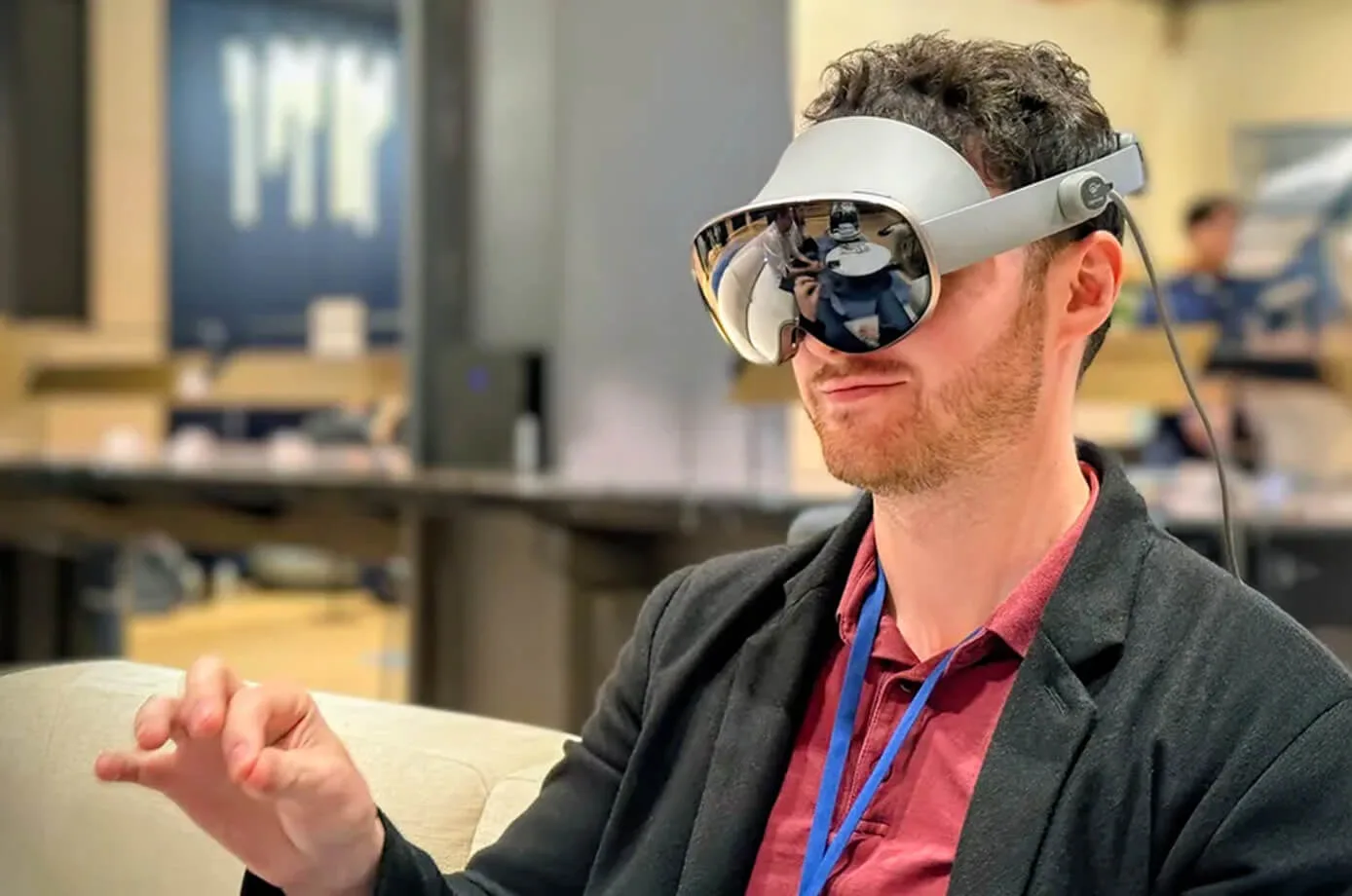 Seated bearded man wearing a silver-grey mixed-reality headset with a trailing cable, raising his right hand in a mid-air pinch gesture
