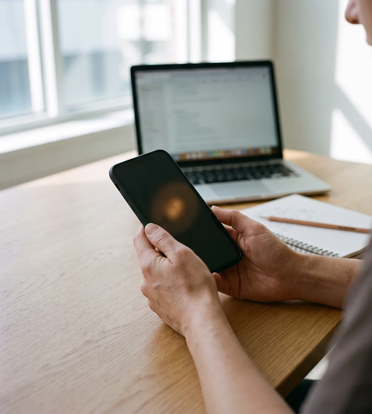 Hands holding an iPhone at a developer’s desk beside an open laptop