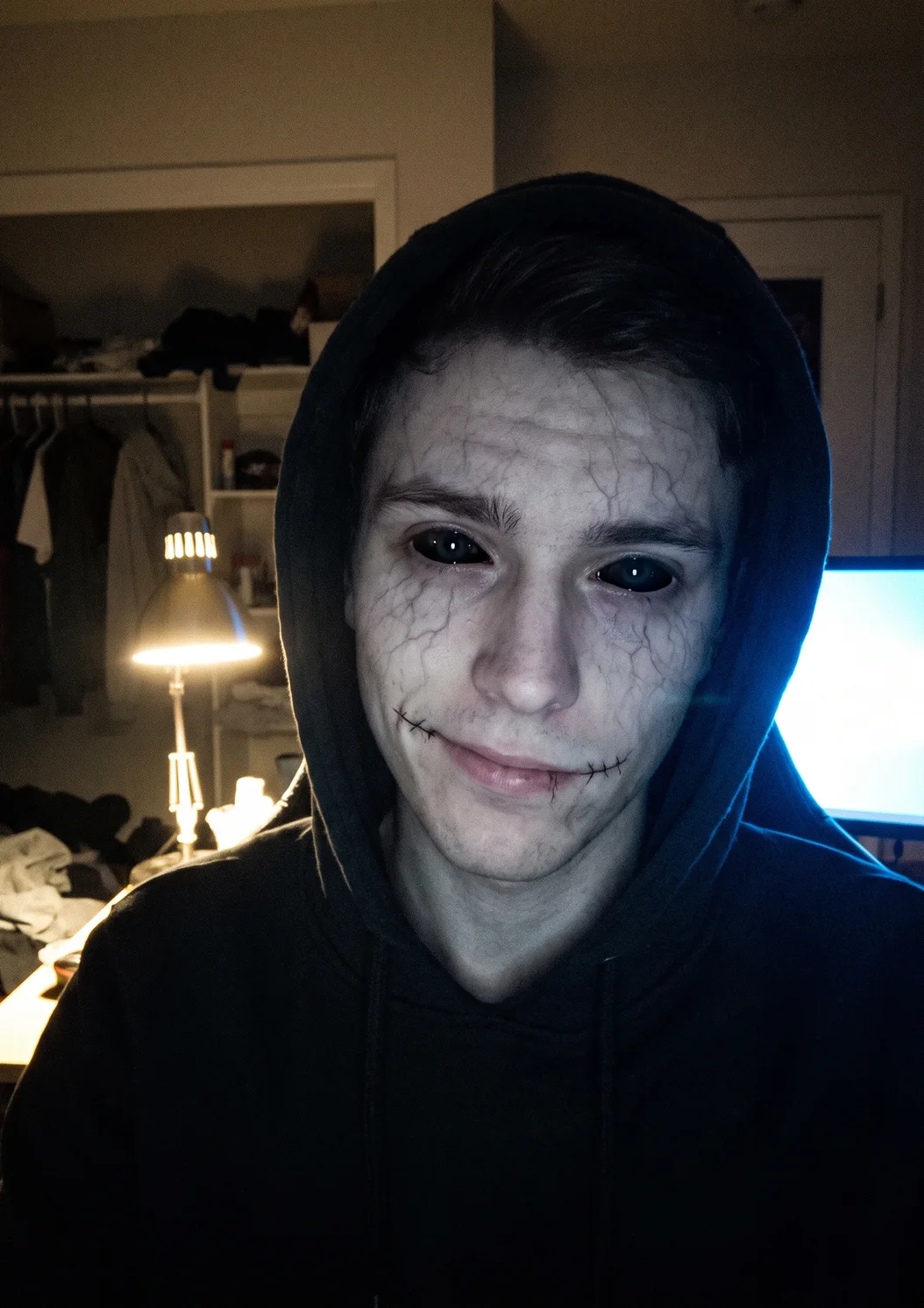 Fan selfie in a hooded sweatshirt using a horror face lens: jet-black eyes, faint grey veins and stitched mouth corners