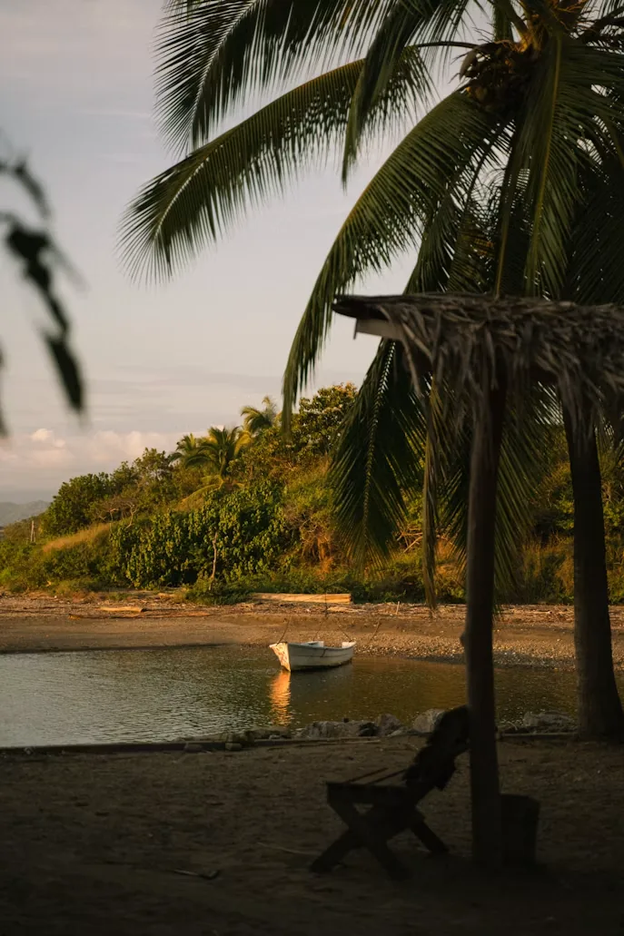 A palm-thatched shade and a rowing boat on a quiet lagoon beach at dusk