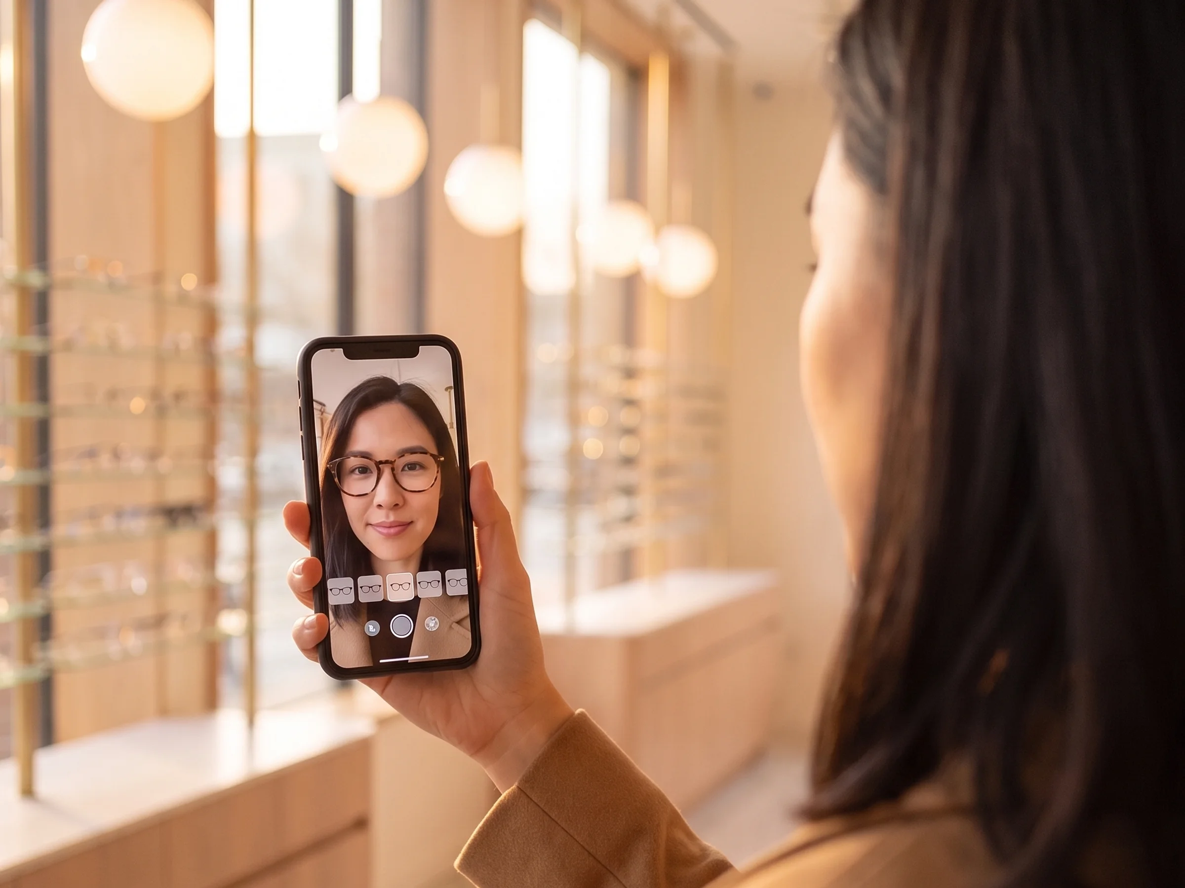 Augmented reality in retail — over-the-shoulder view of a shopper in an eyewear boutique holding up her phone, where a virtual try-on shows tortoiseshell frames on her face
